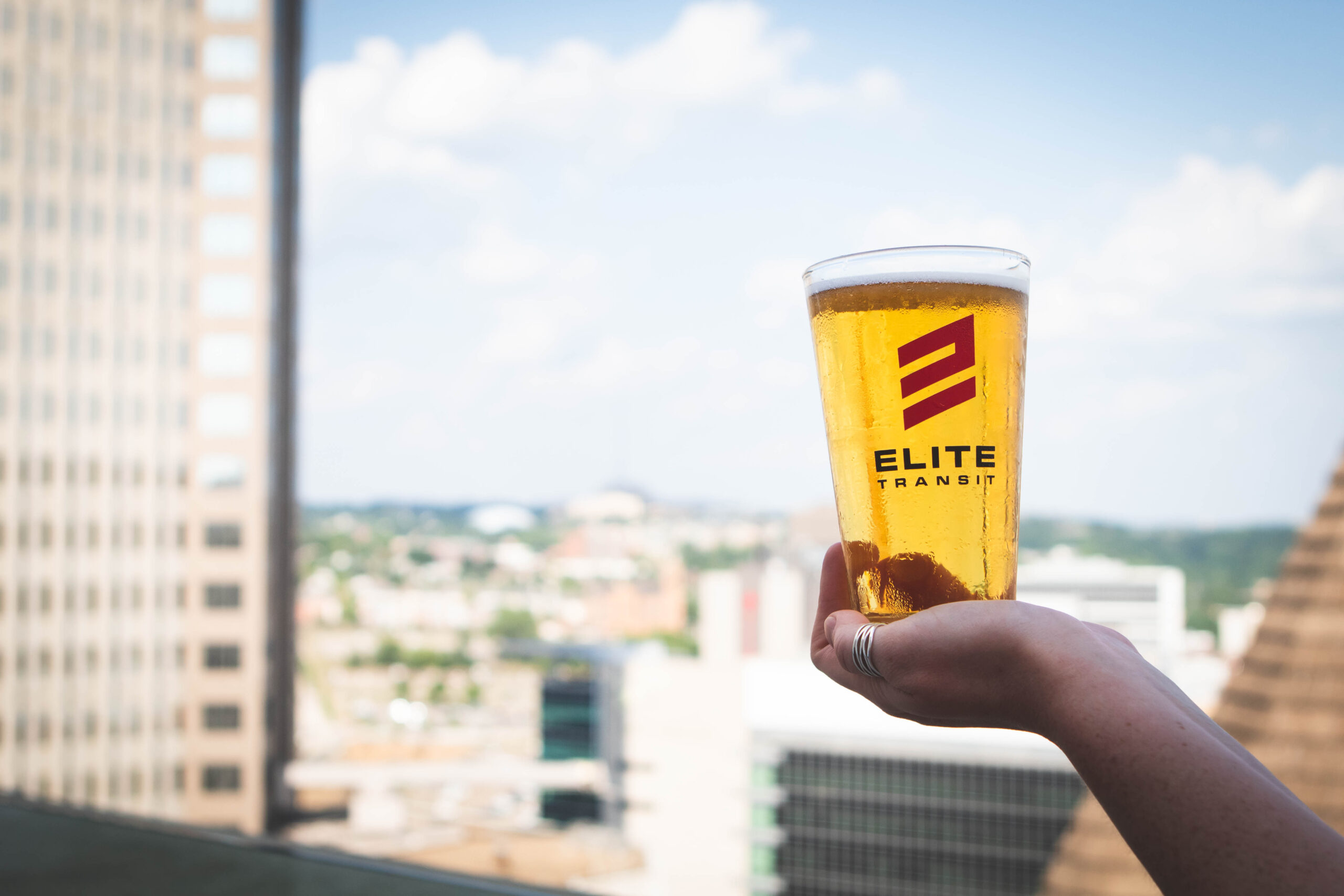 Pale beer pint glass in ELITE Transit Solutions glassware on the balcony with downtown Pittsburgh out of focus in the background.
