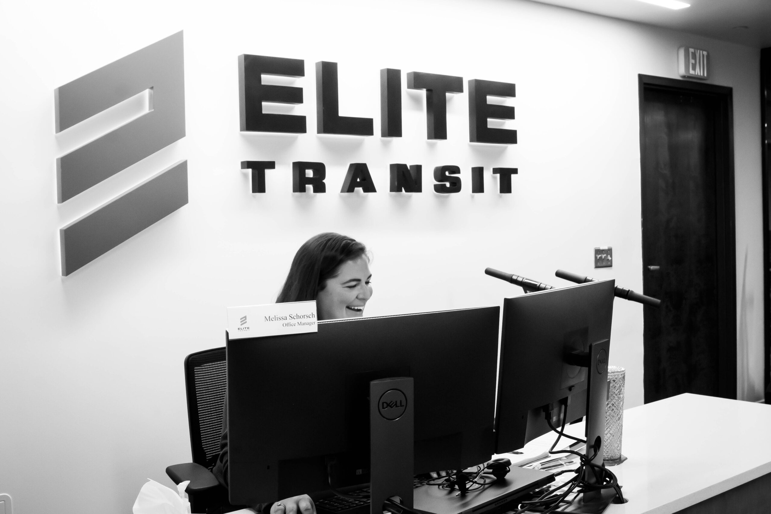 Black and white photo of ELITE Transit Solutions Charlotte office greeting area with friendly office manager and ELITE Transit Solutions logo on the wall.
