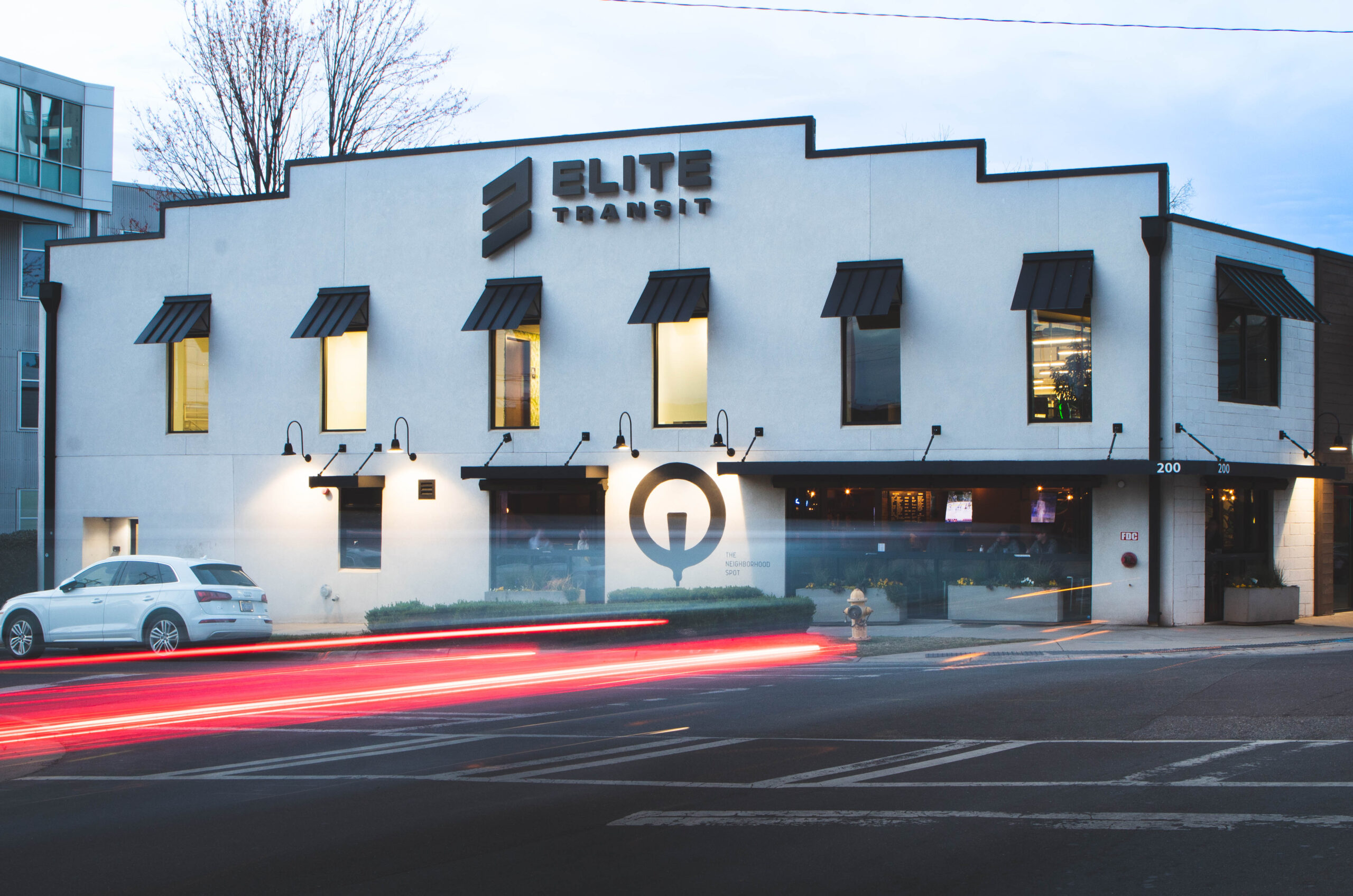 ELITE Transit Solutions exterior in Charlotte, NC. It's a white building with black shutters above QC Pourhouse, a popular bar with a black logo. The exposure is delayed causing a red blur from car headlights.